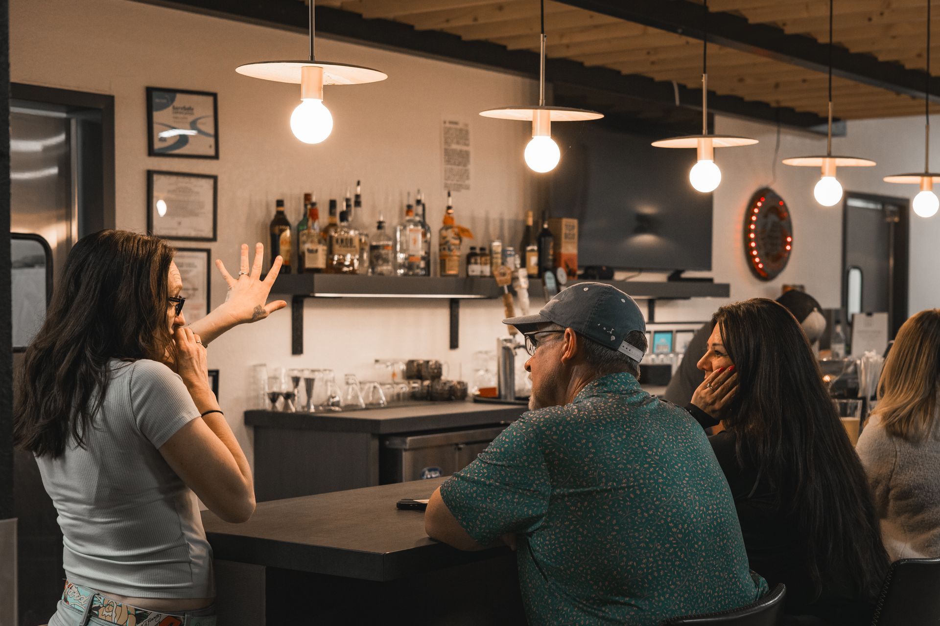 People chatting at a bar counter under warm pendant lights, with bottles and decor behind them.