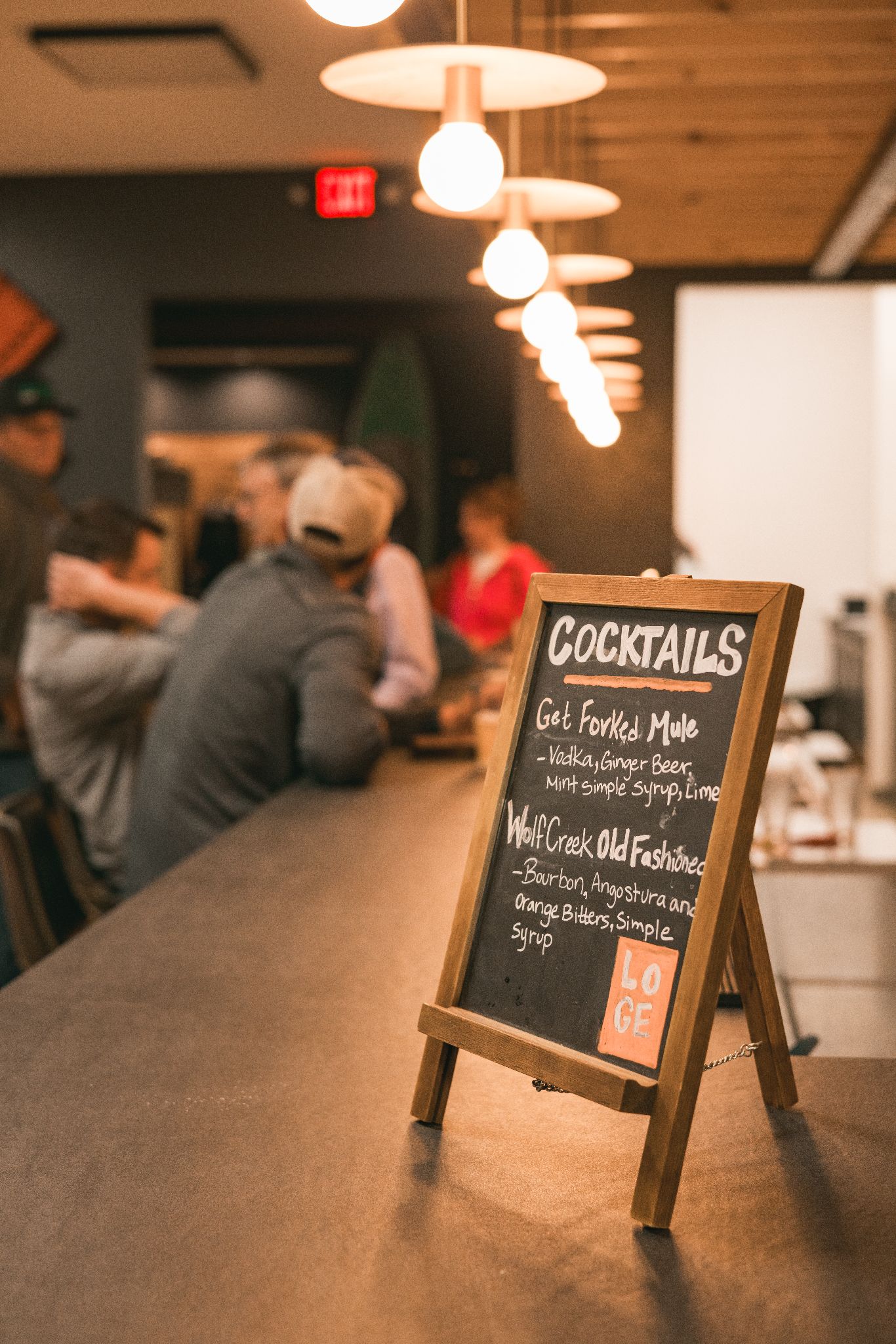 Cocktail menu on a bar counter with blurred patrons and hanging lights in the background