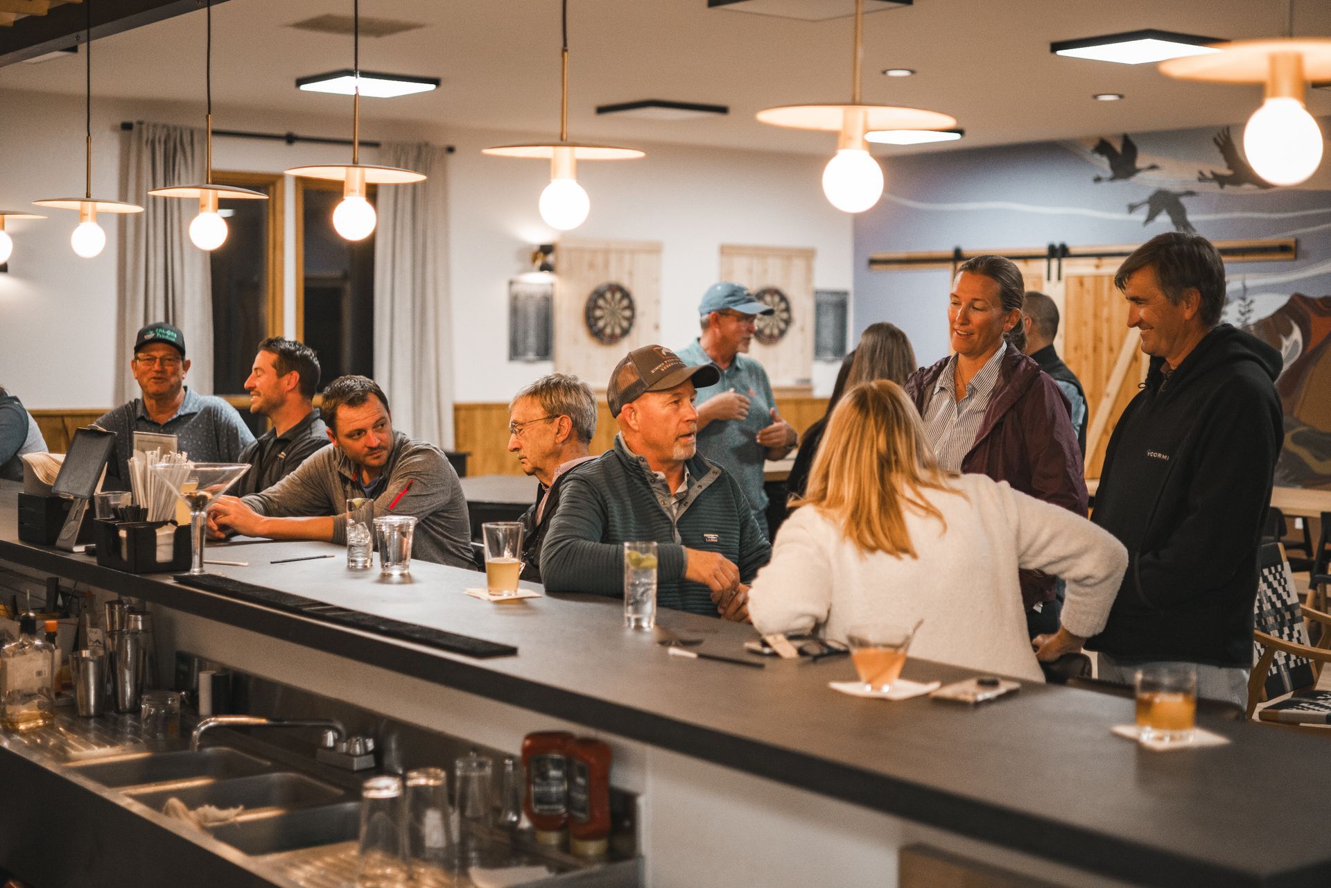 People chatting and drinking at a cozy bar with warm hanging lights and a long counter