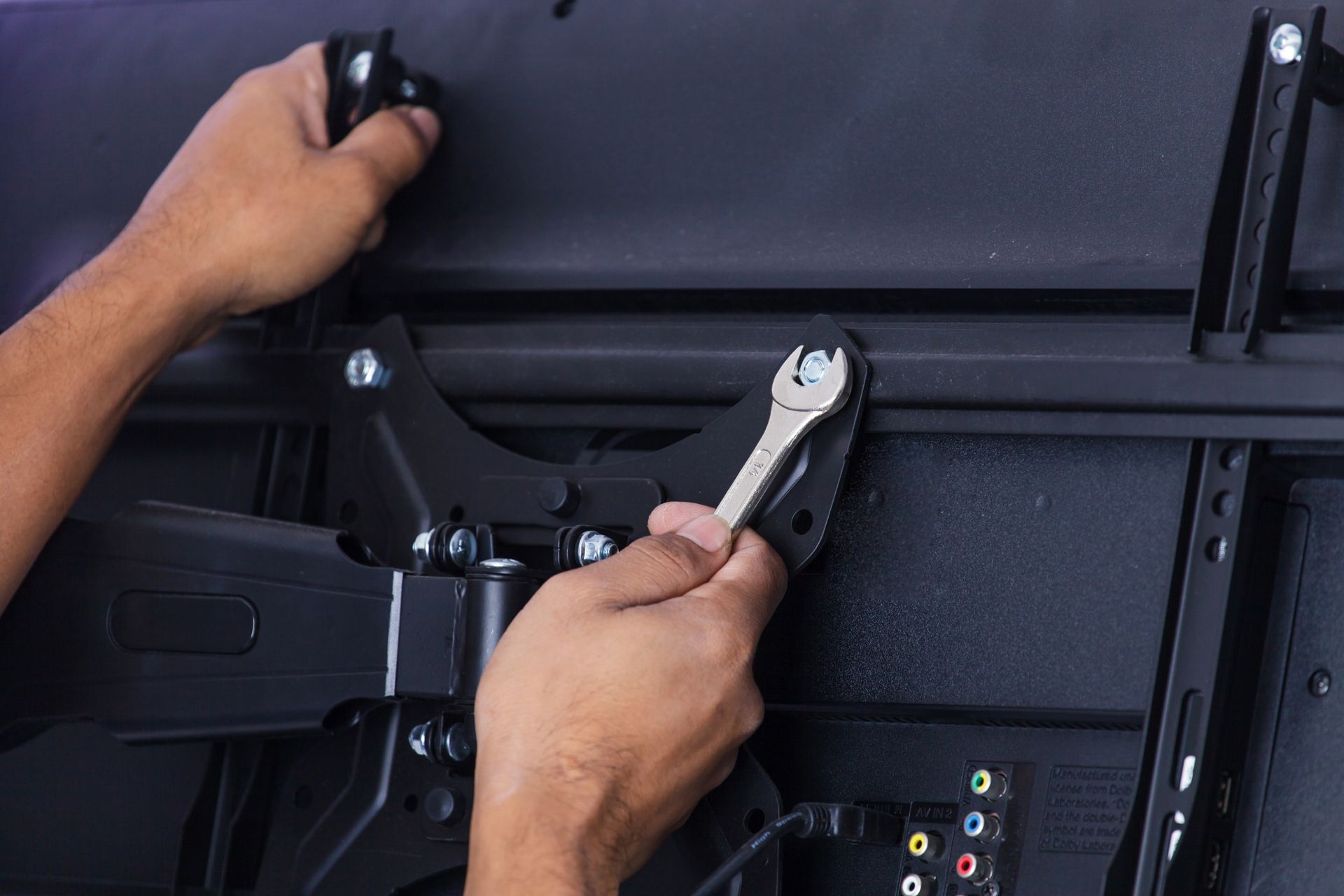 Hands using a wrench to install a black wall-mounted TV bracket on a flat-screen TV.