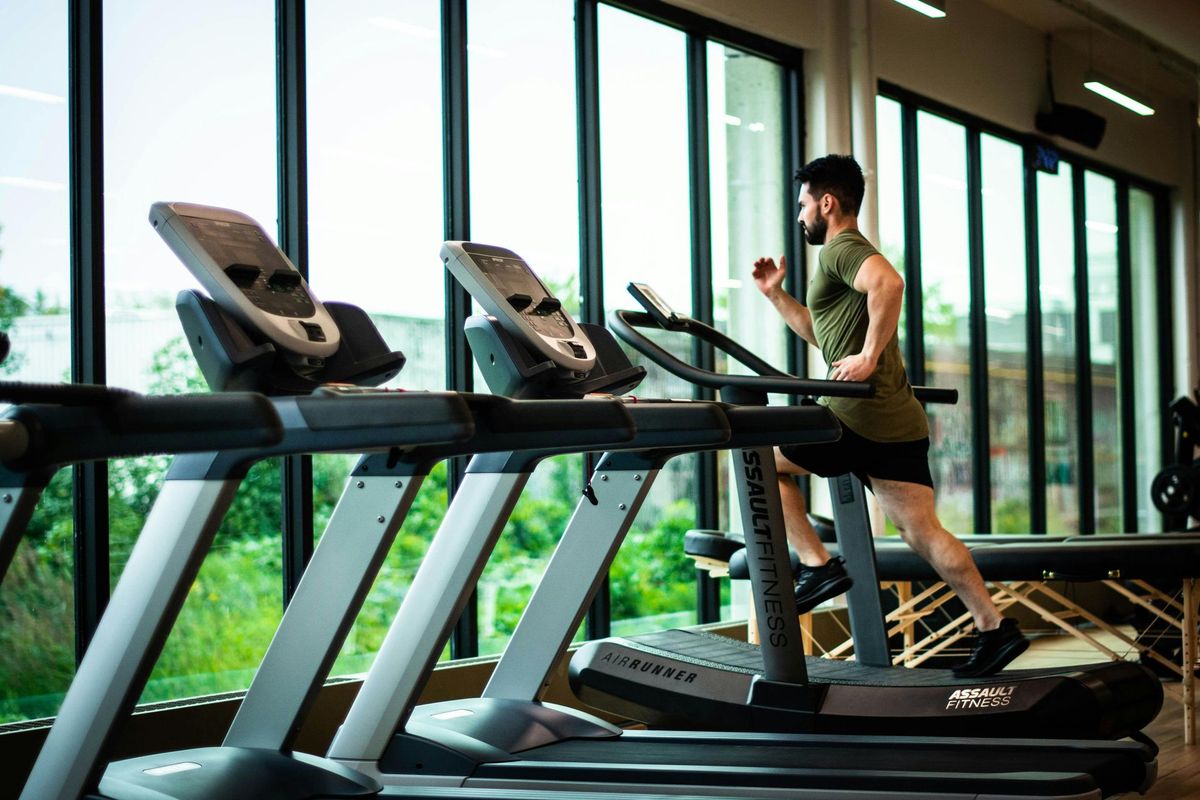 Man running on a treadmill in a gym, with large windows overlooking green foliage.
