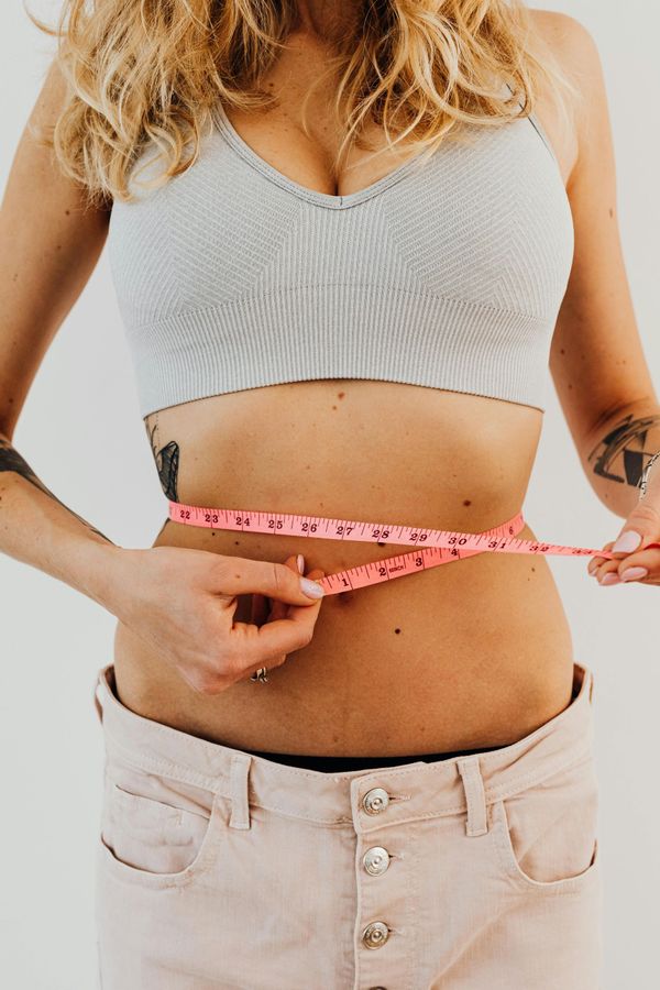 Woman in a crop top and pants measures waist with a pink tape measure.