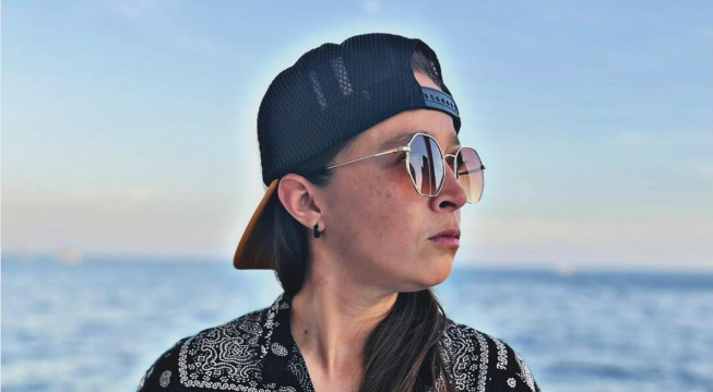 Persona con gafas de sol y gorra hacia atrás mirando hacia un lado con un fondo de océano y cielo.