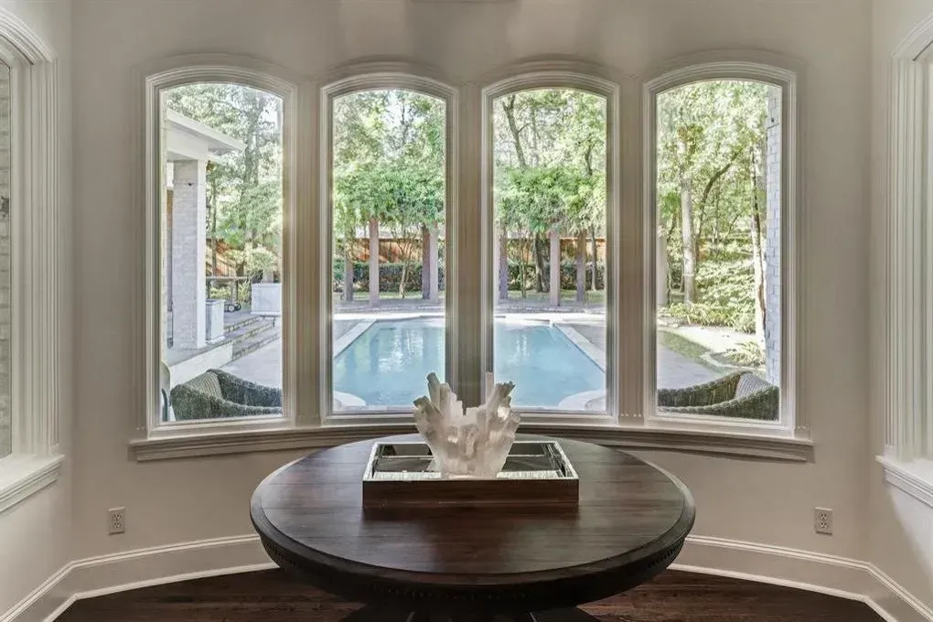 A round wooden table with decor in front of arched windows overlooking a swimming pool and yard.