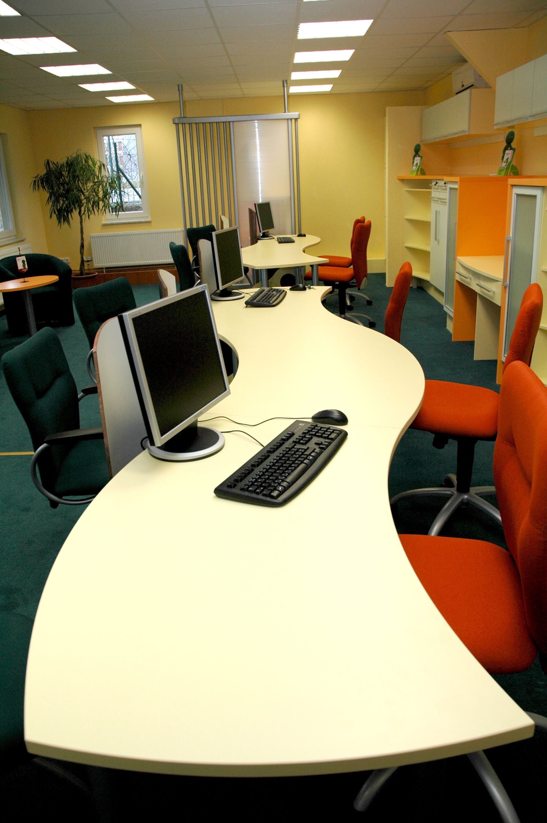 Bureau avec des ordinateurs sur une longue table incurvée ; les chaises sont orange et vertes, et les murs sont jaunes.