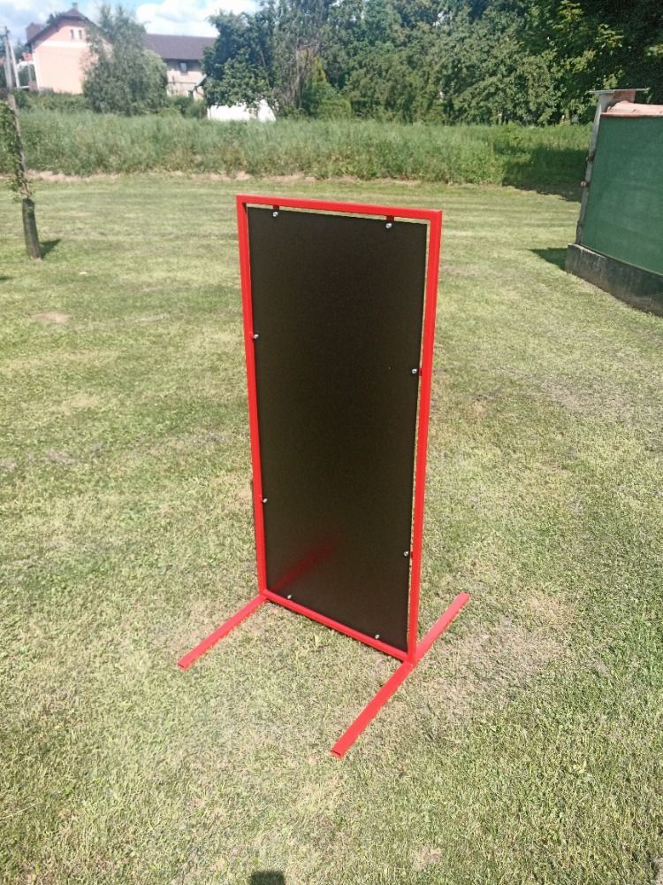 Cible noire à cadre rouge sur un support dans un champ herbeux.
