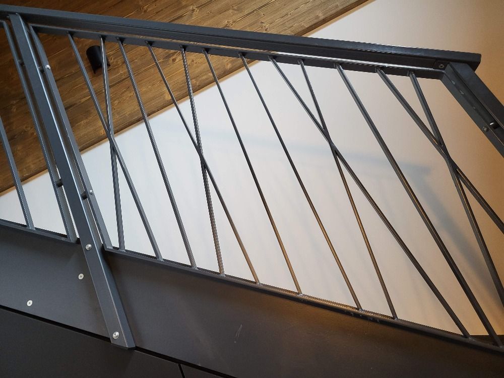 Rambarde d'escalier en métal gris avec barreaux torsadés en diagonale, sur un mur blanc et un plafond en bois.