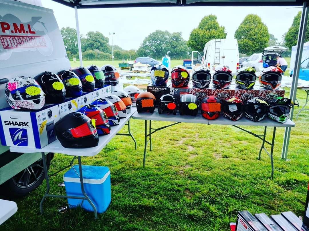 Motorcycle helmets displayed on a table at an outdoor event, with grass and parked vehicles visible.