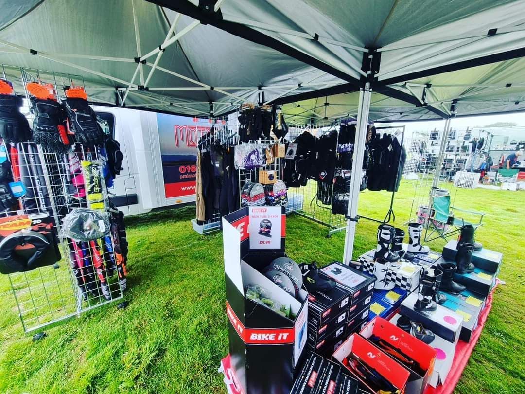 Market stall with outdoor gear under a white canopy on a grassy field; products include backpacks and accessories.