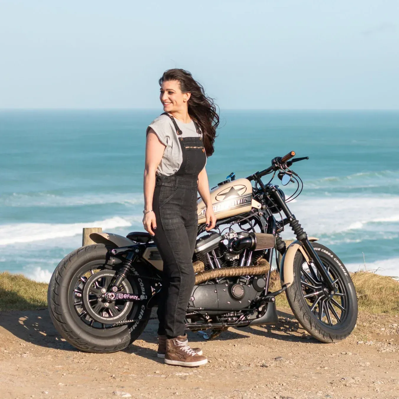 Woman in overalls stands next to a beige motorcycle overlooking ocean.