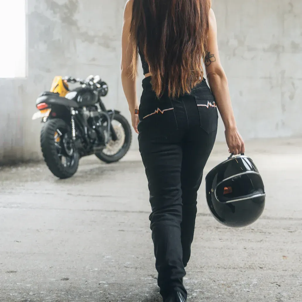 Woman in black attire walks away from motorcycle, holding a helmet, indoors.