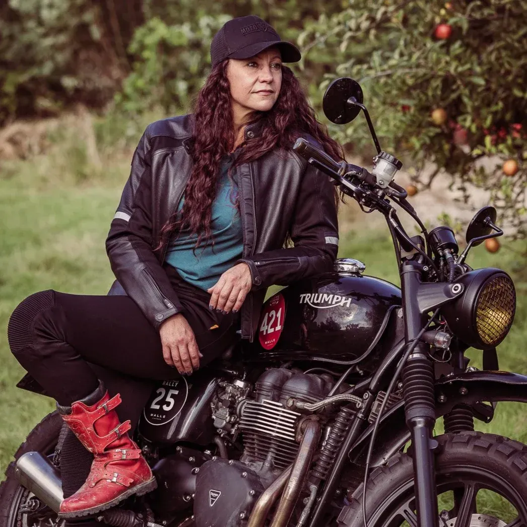 Woman in motorcycle gear sits on a black Triumph bike, red boots, looking to the side in a garden.