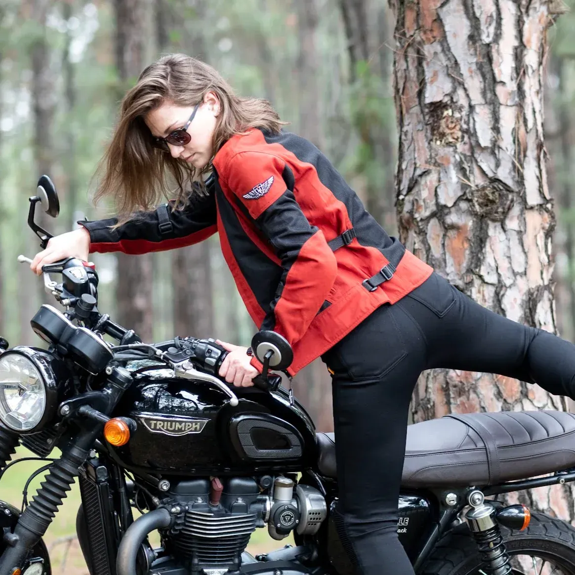 Woman in red motorcycle jacket, black pants leaning on black Triumph motorcycle in wooded area.