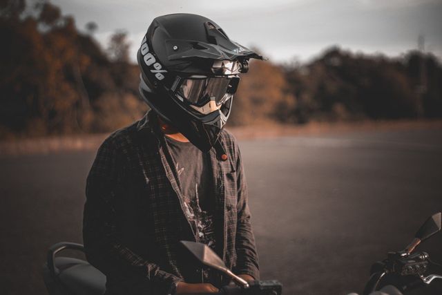 Person wearing a black motorcycle helmet and jacket stands near a motorcycle, outdoors.