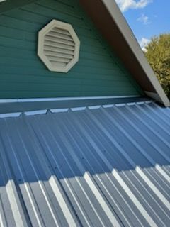 Metal roof with blue-green siding, octagonal vent, and brown trim against a partly cloudy sky.