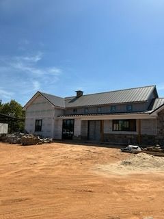 A house under construction with a metal roof and stone foundation on a dirt lot under a blue sky.