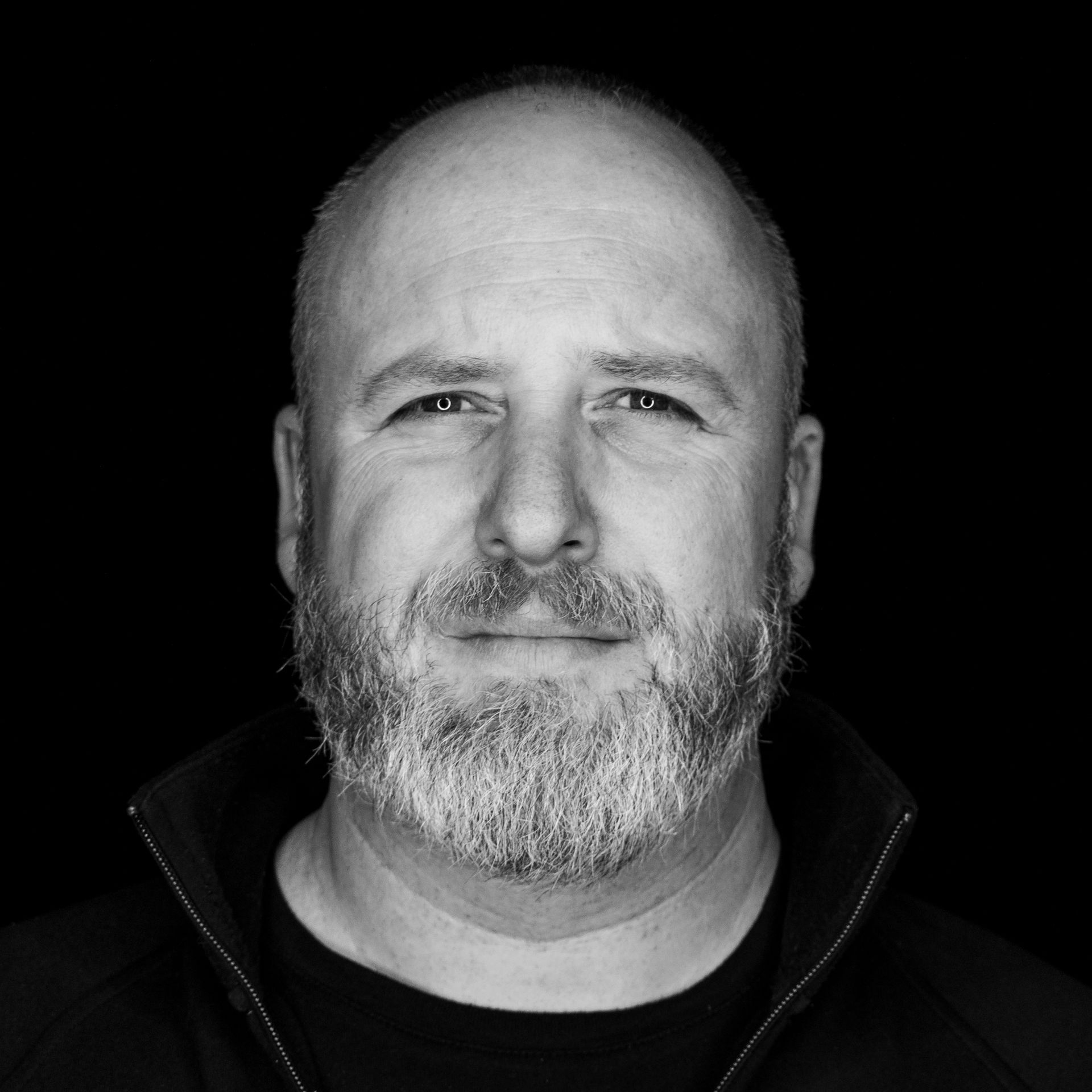 Man with a salt-and-pepper beard looks directly at the camera, black and white portrait, dark background.