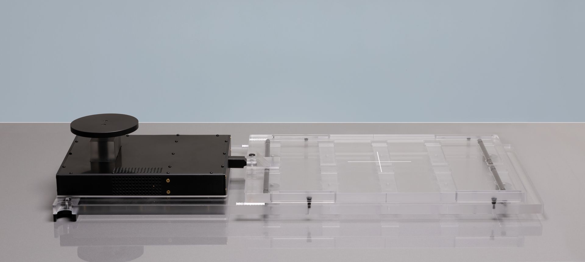 A black device with a round knob sits on a clear plastic tray. It is on a gray surface against a light blue background.