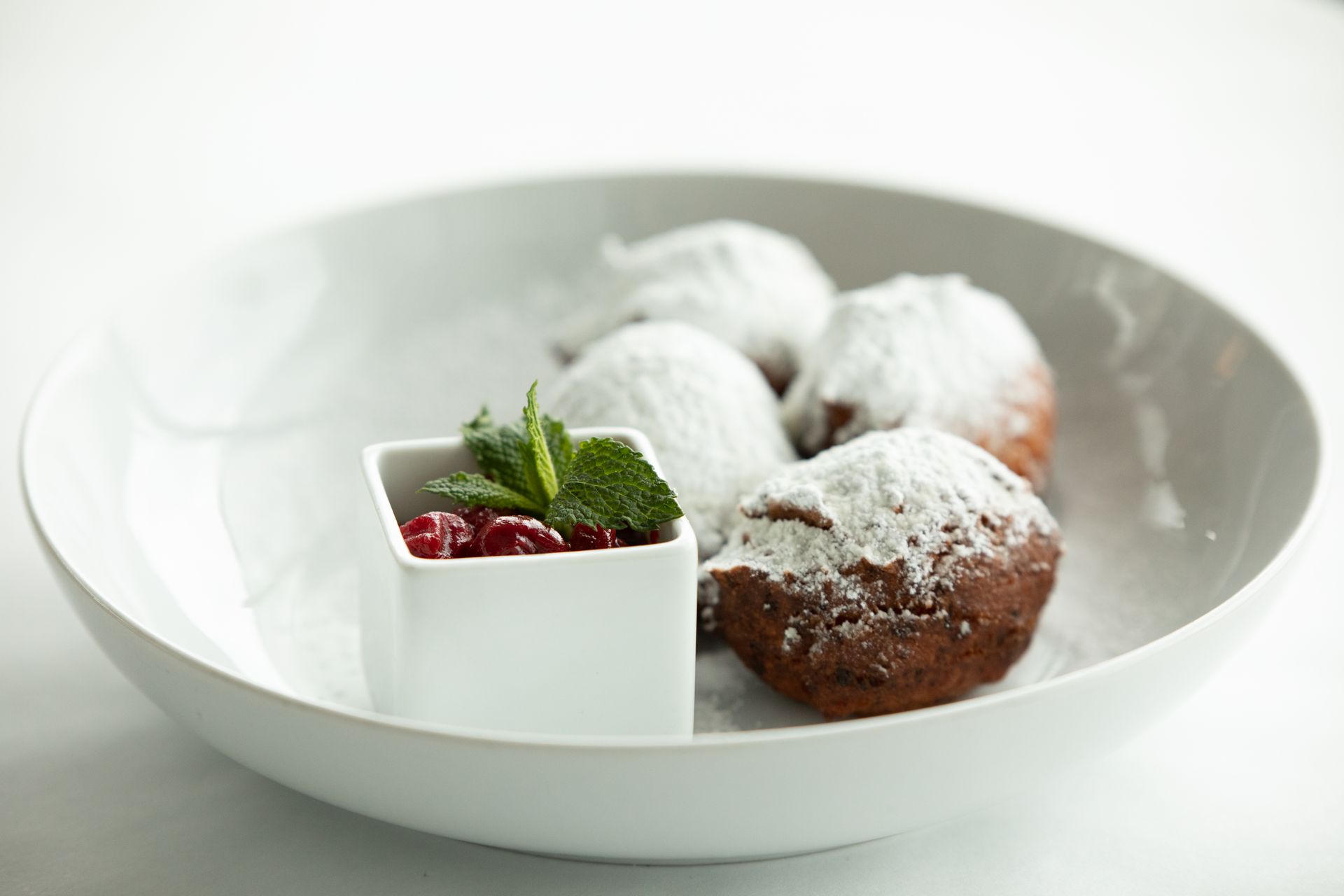 Beignets dusted with powdered sugar, served with a side of red jam and mint in a white bowl.