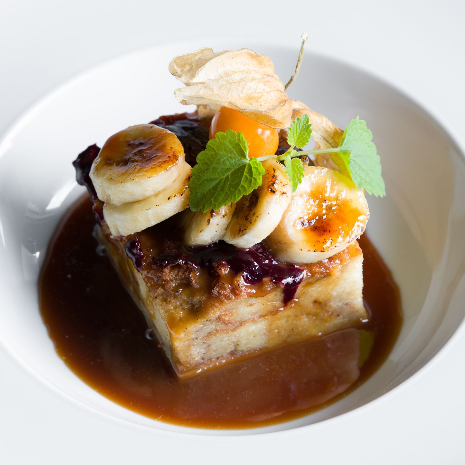 Bread pudding with caramelized bananas, berry compote, and sauce in a white bowl.