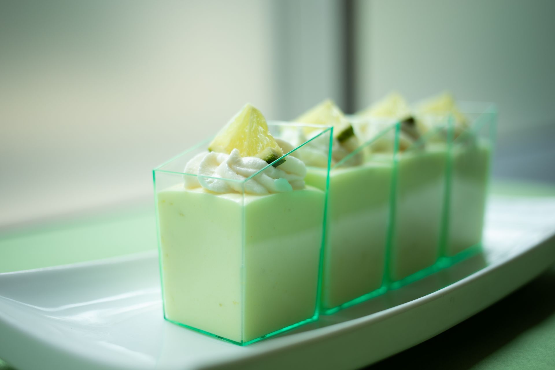 Green layered dessert in clear cups, topped with whipped cream and citrus, served on a white rectangular plate.