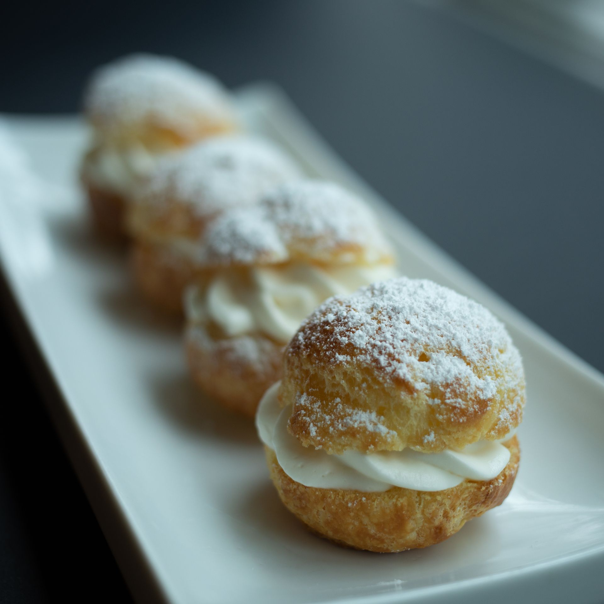 Cream puffs, filled with white cream, dusted with powdered sugar, arranged on a white plate.