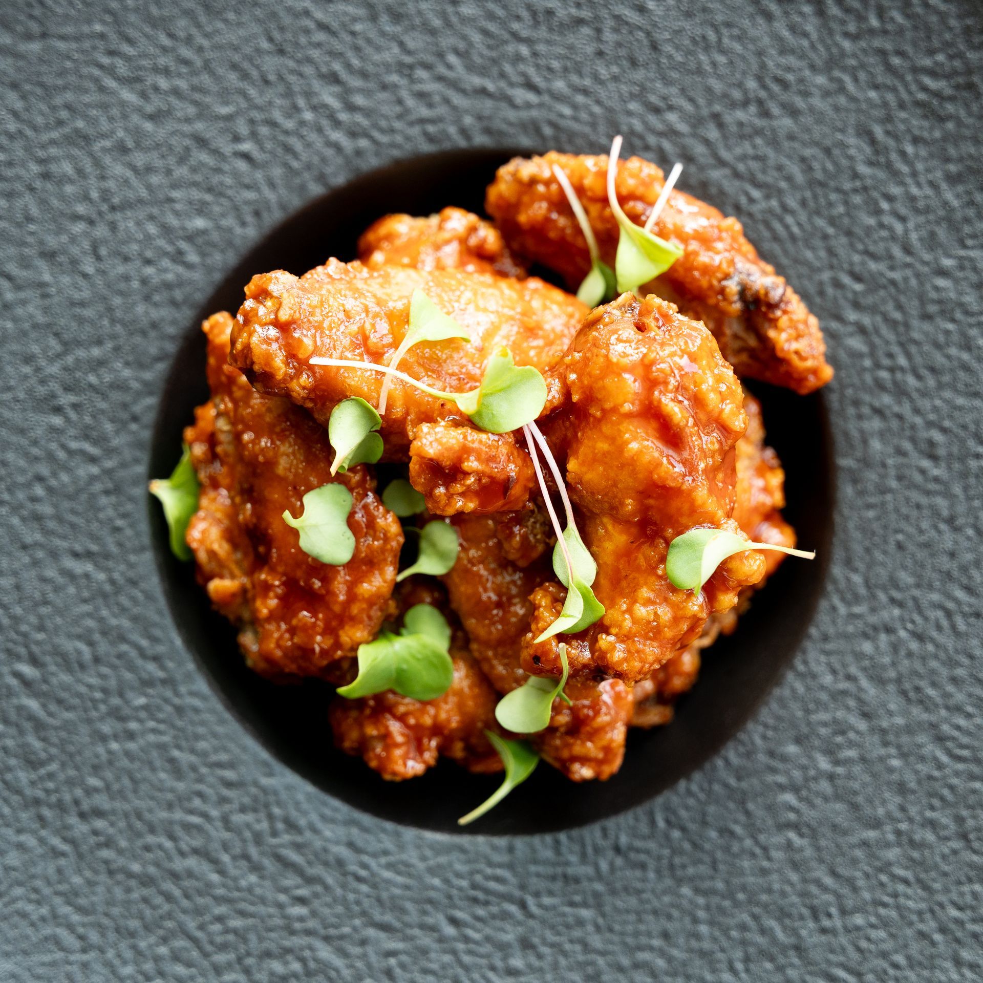 Fried chicken wings with microgreens in a black bowl on a gray surface.