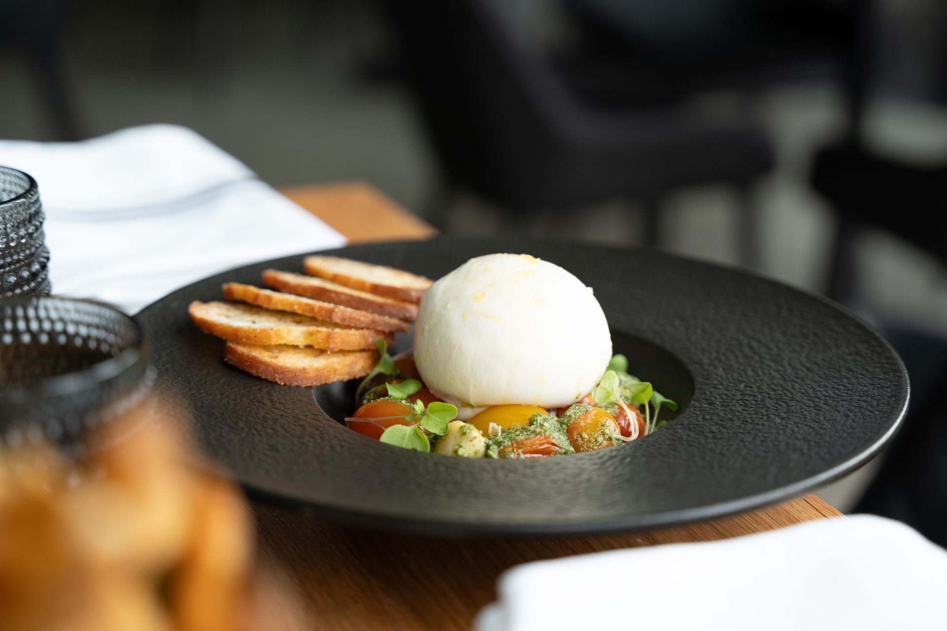 Burrata cheese ball on a bed of greens and tomatoes, with toasted bread, served on a black plate.