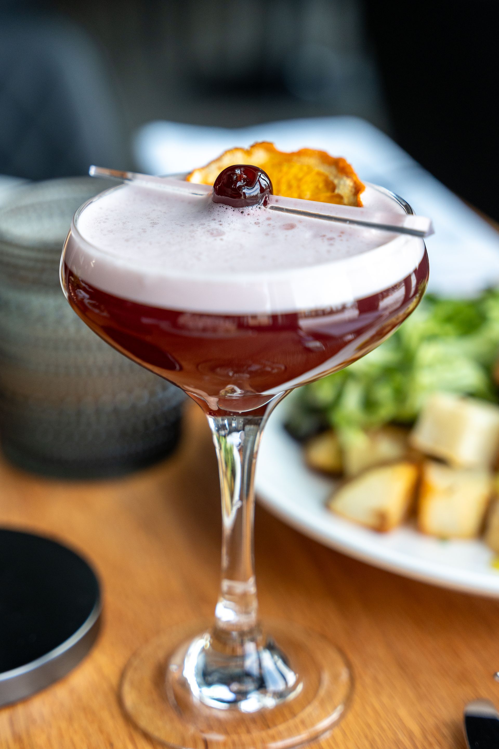 Cocktail in a stemmed glass, topped with foam and a cherry garnish, on a wooden table.