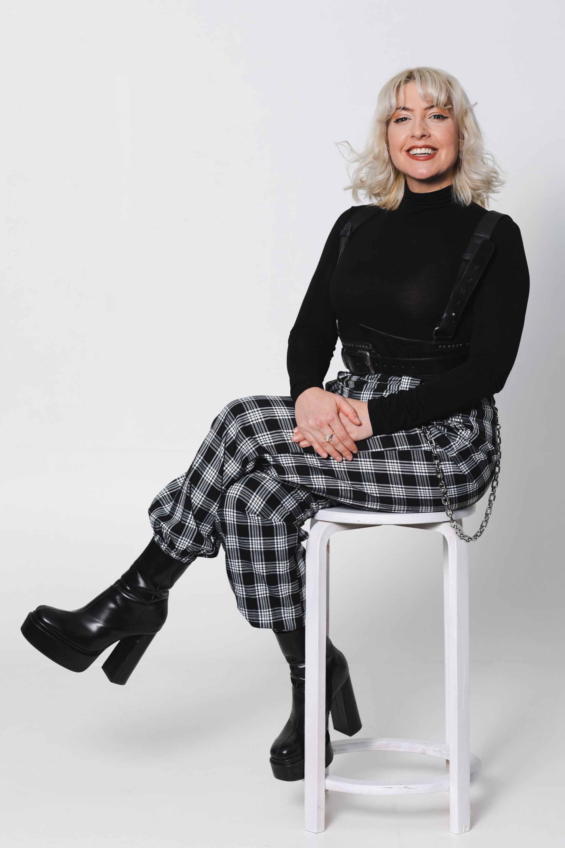 Woman in black turtleneck and plaid pants, seated on a stool, smiling, and wearing platform boots.