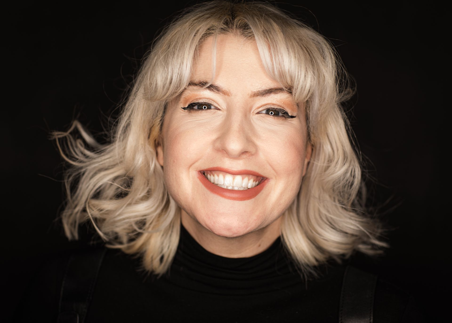Woman with blonde wavy hair, smiling, wearing black turtleneck and eyeliner, against a black background.