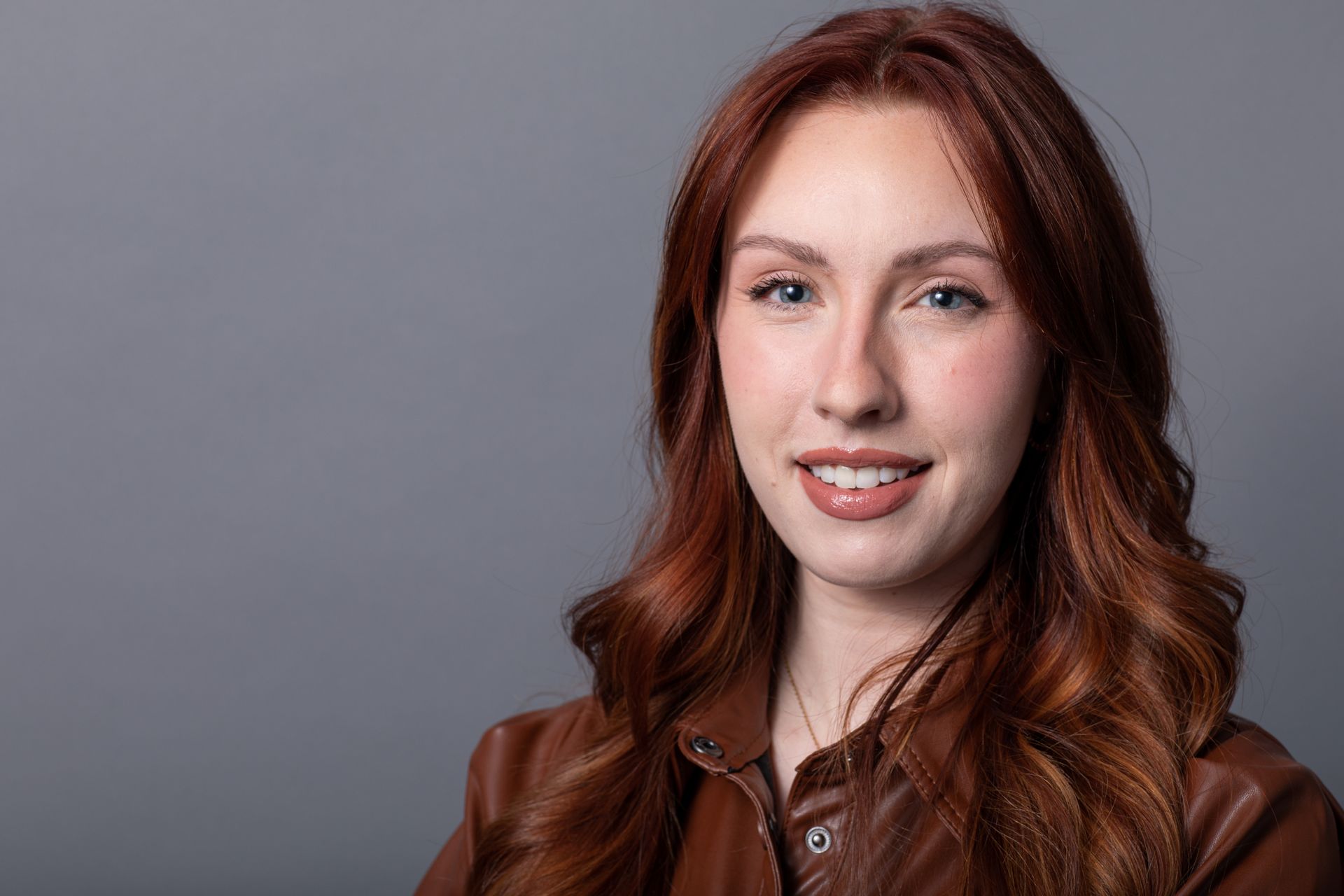 Woman with red wavy hair smiles in a brown leather jacket against a gray backdrop.