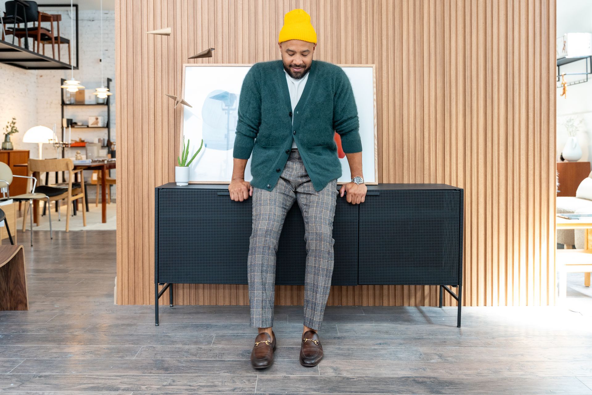 Man in patterned pants, green cardigan, and yellow hat stands in front of a wood-paneled wall and black cabinet.