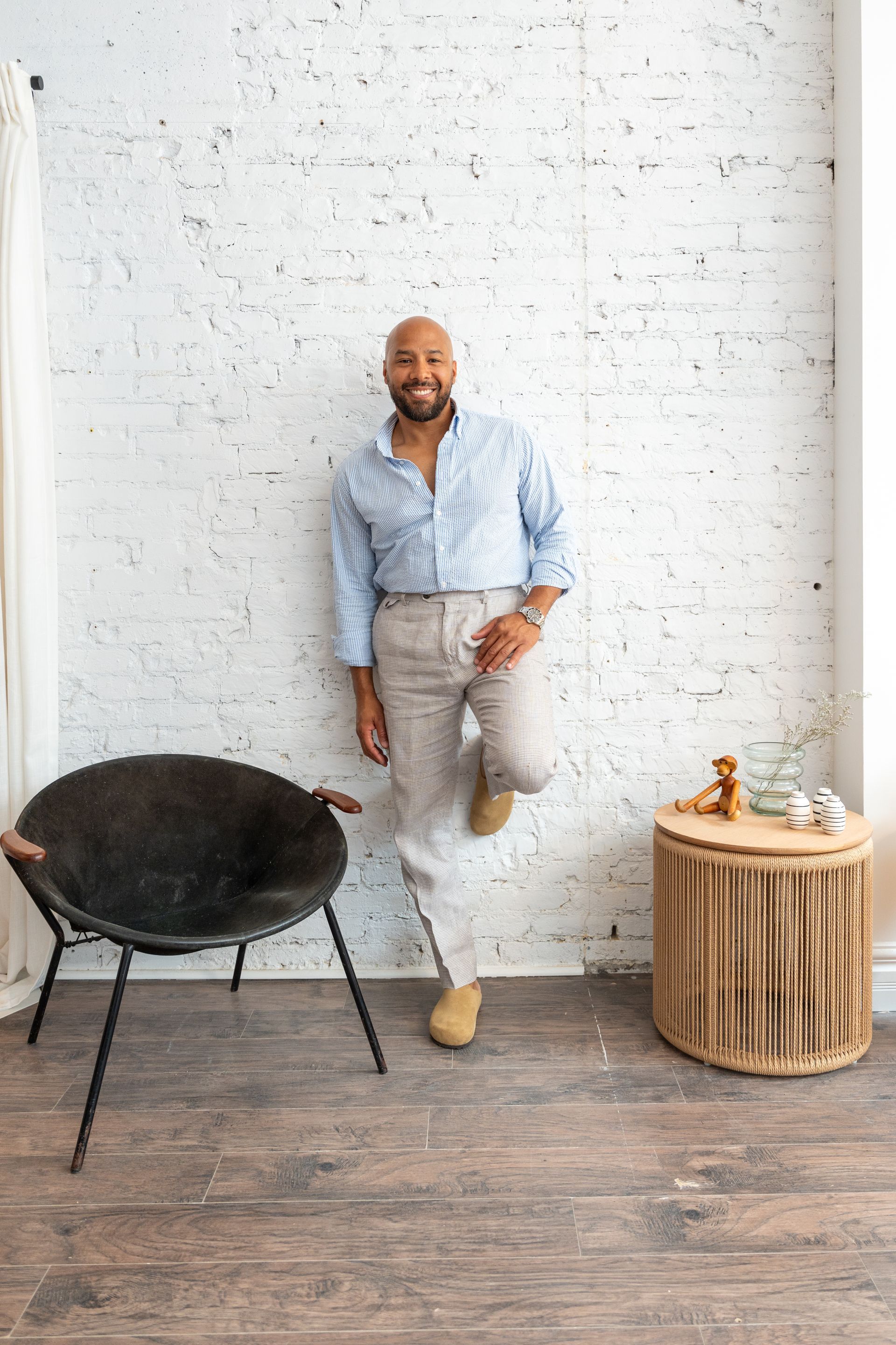Man leans against white brick wall, wearing patterned shirt and pants. Black chair and side table nearby.