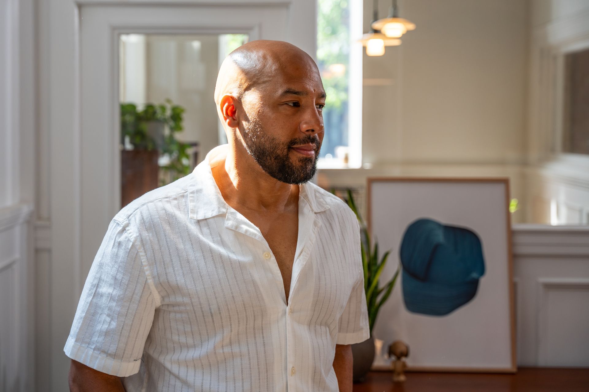 Man with shaved head and beard in a white shirt, looking to the side in a well-lit room.