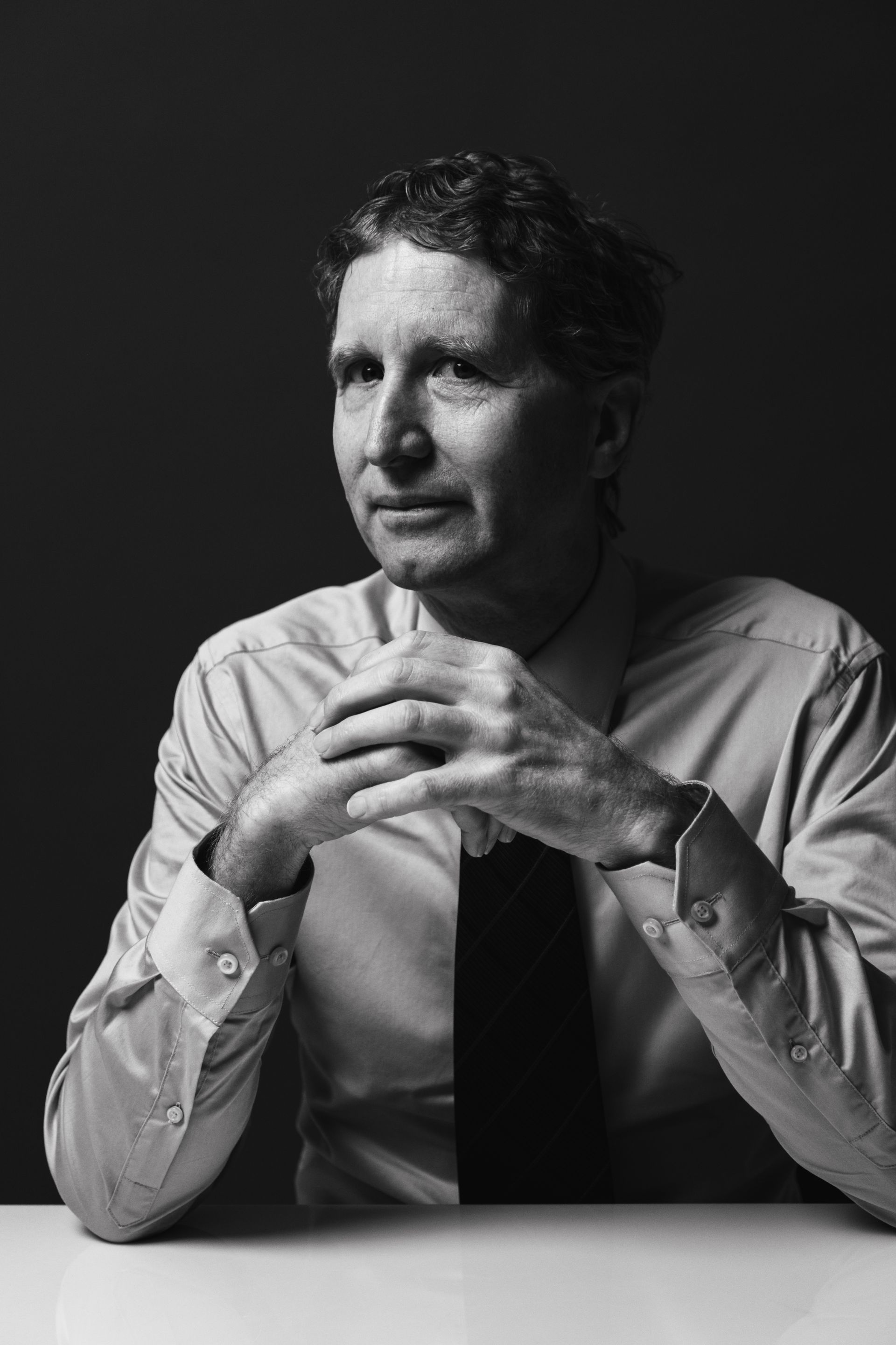 Man in a light-colored shirt and tie, arms crossed, sits at a table, looking to the side.