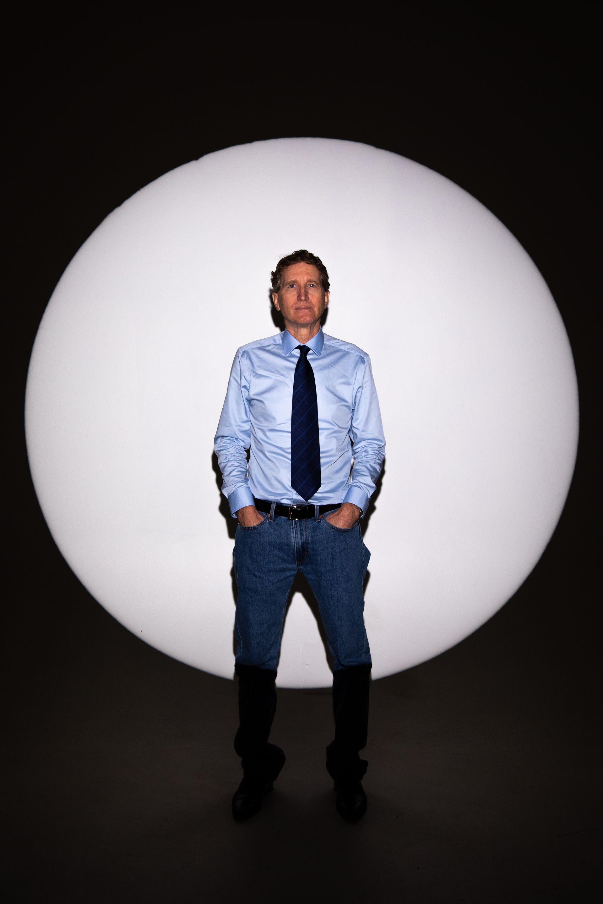 Man in button-down shirt and tie stands with hands in pockets before a large white orb.