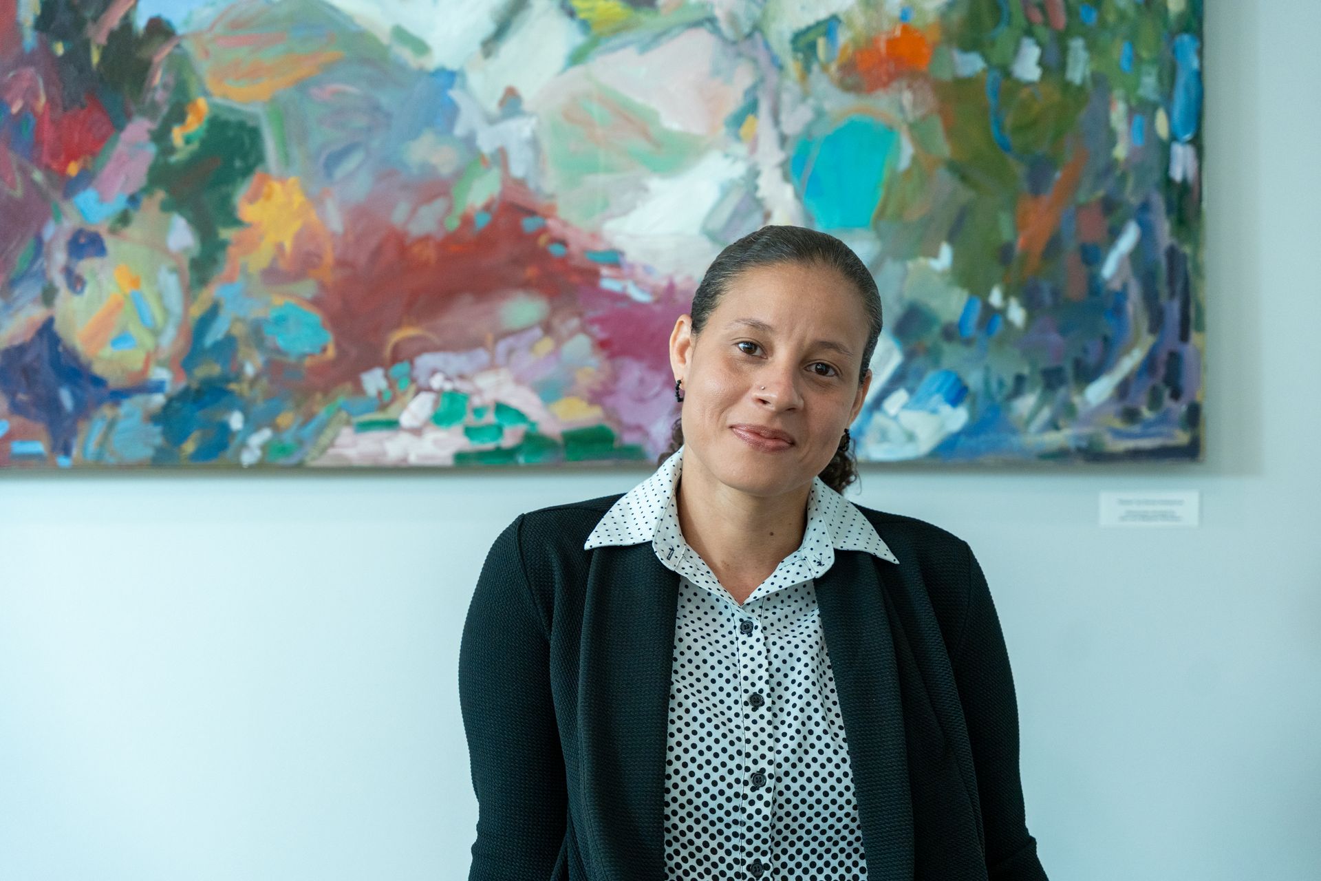 Woman in black jacket and patterned shirt smiles in front of colorful abstract painting.