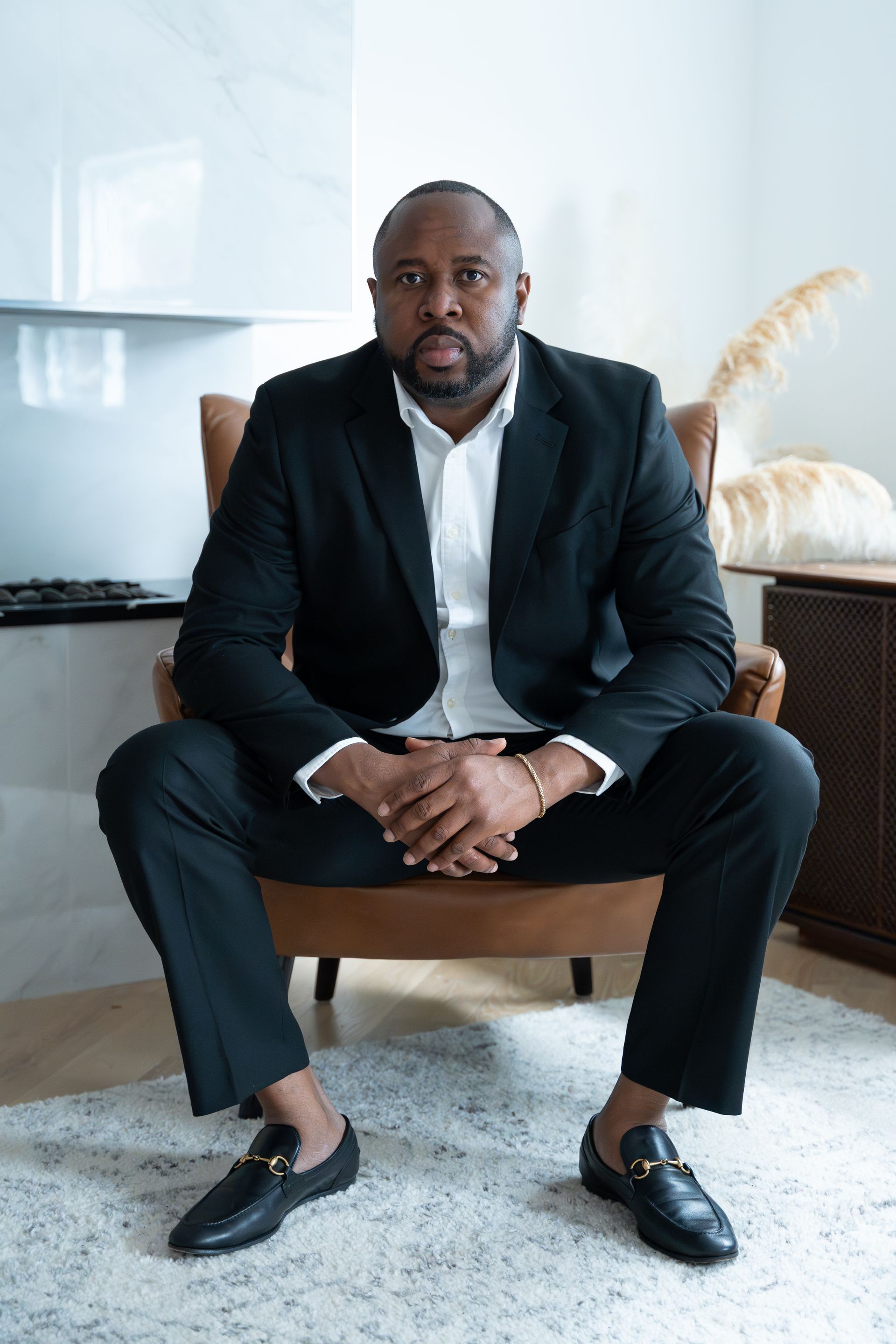 Man in black suit, seated, hands clasped, looking at the camera.