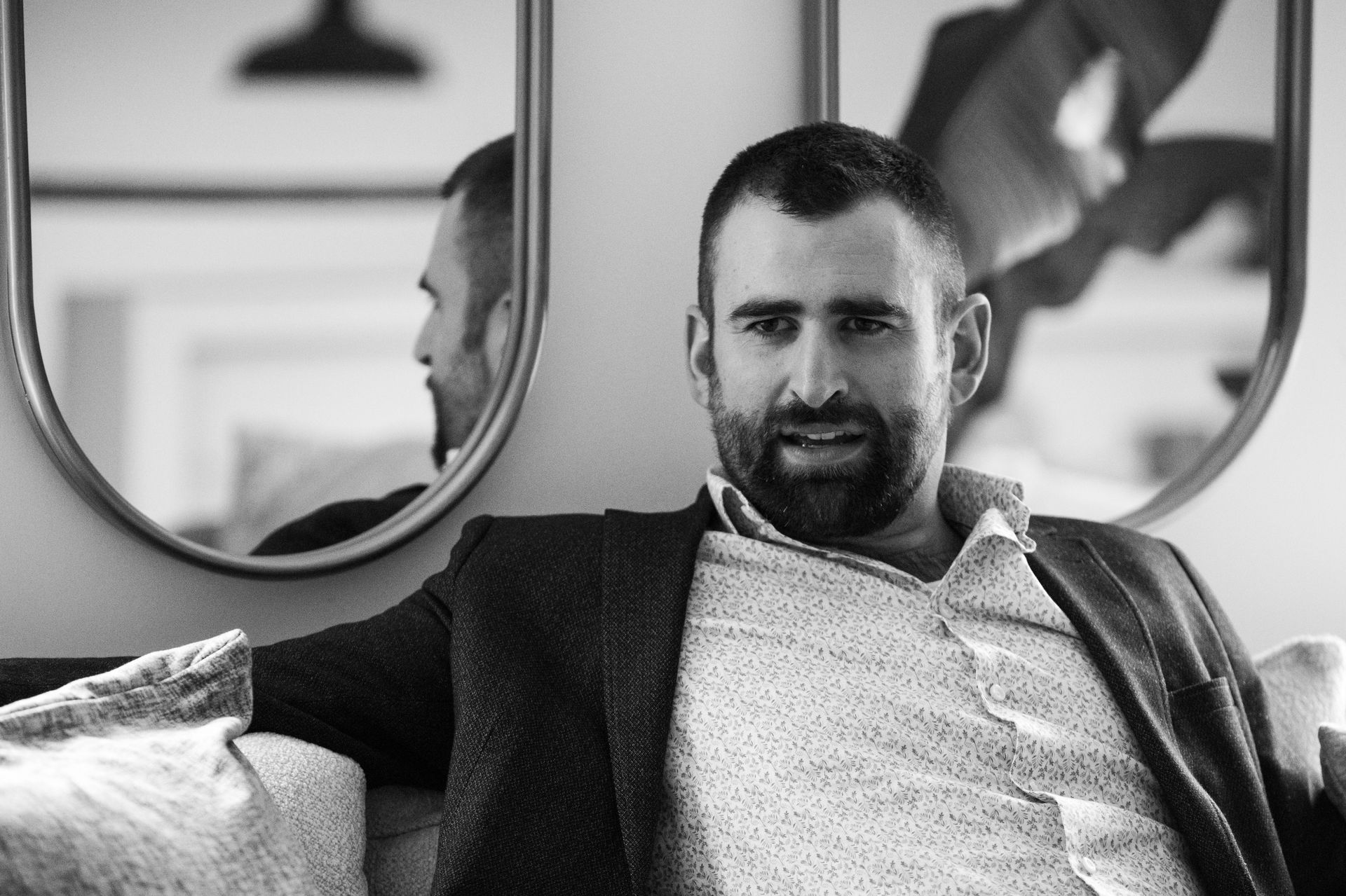Man in blazer and patterned shirt sits on a couch, near a mirror.