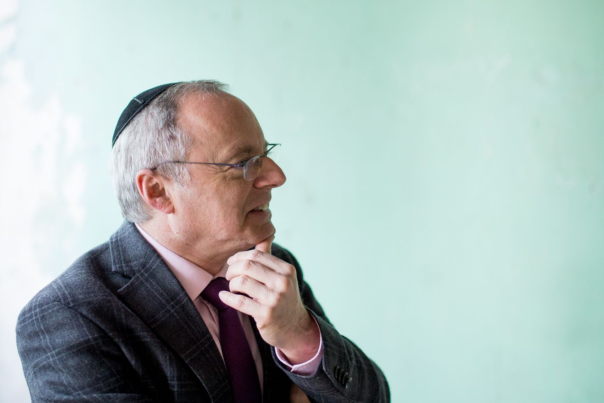 Man in suit and kippah, hand on chin, looking to the side, light green background.