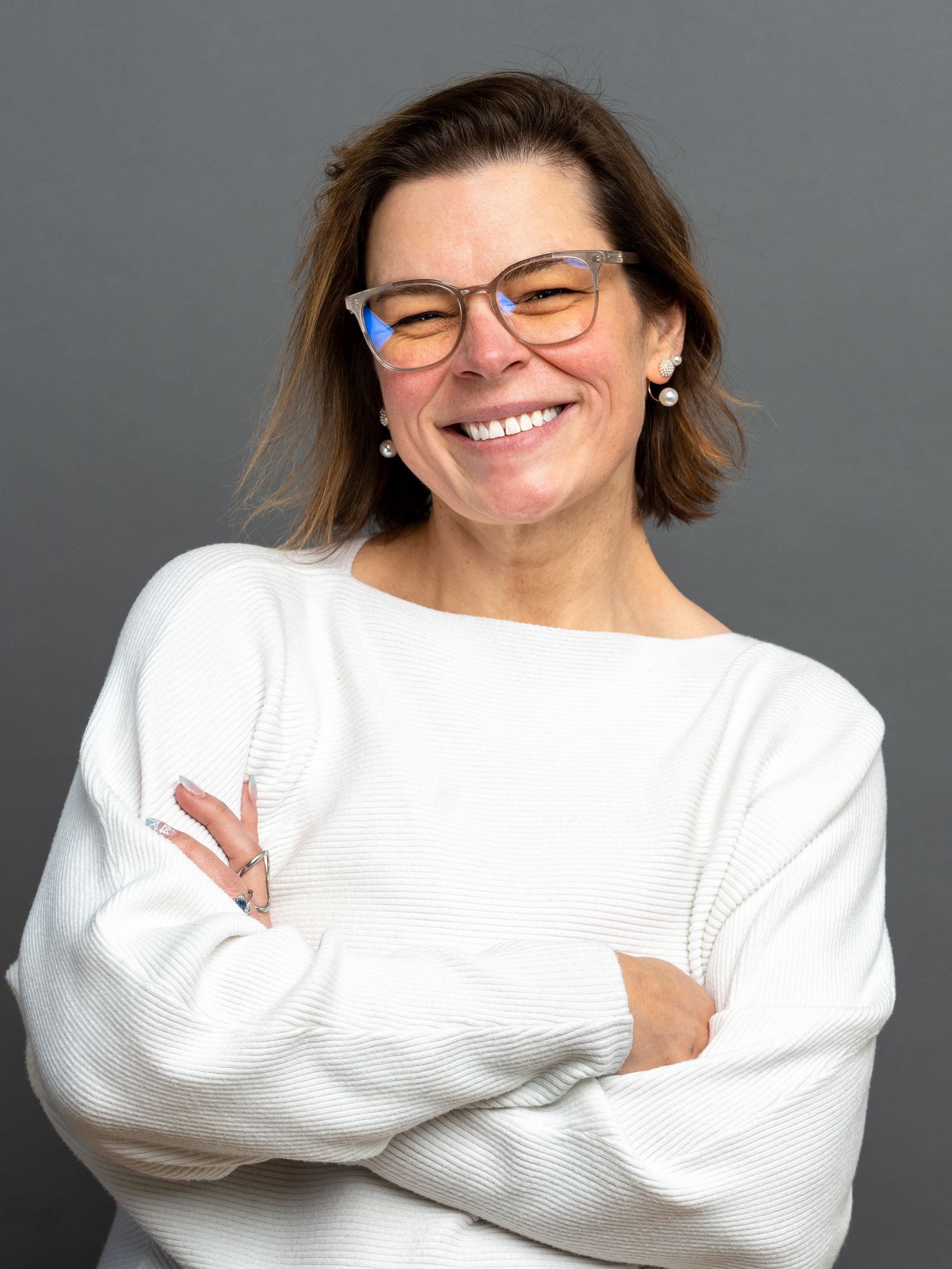 Woman wearing glasses, smiling, arms crossed. She is wearing a white sweater, and the background is gray.