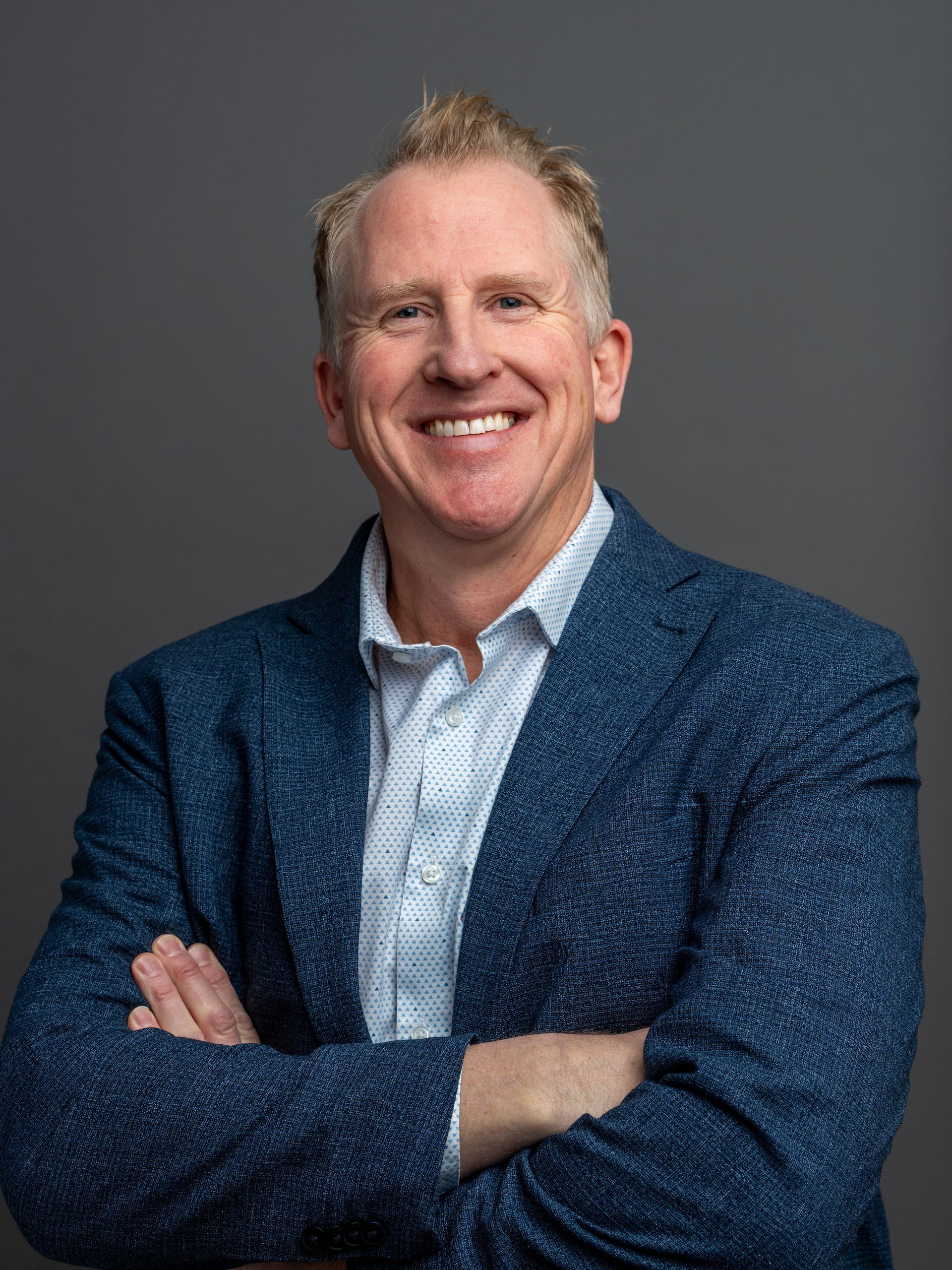Man with crossed arms smiling, wearing a blue blazer over a patterned shirt, against a gray background.