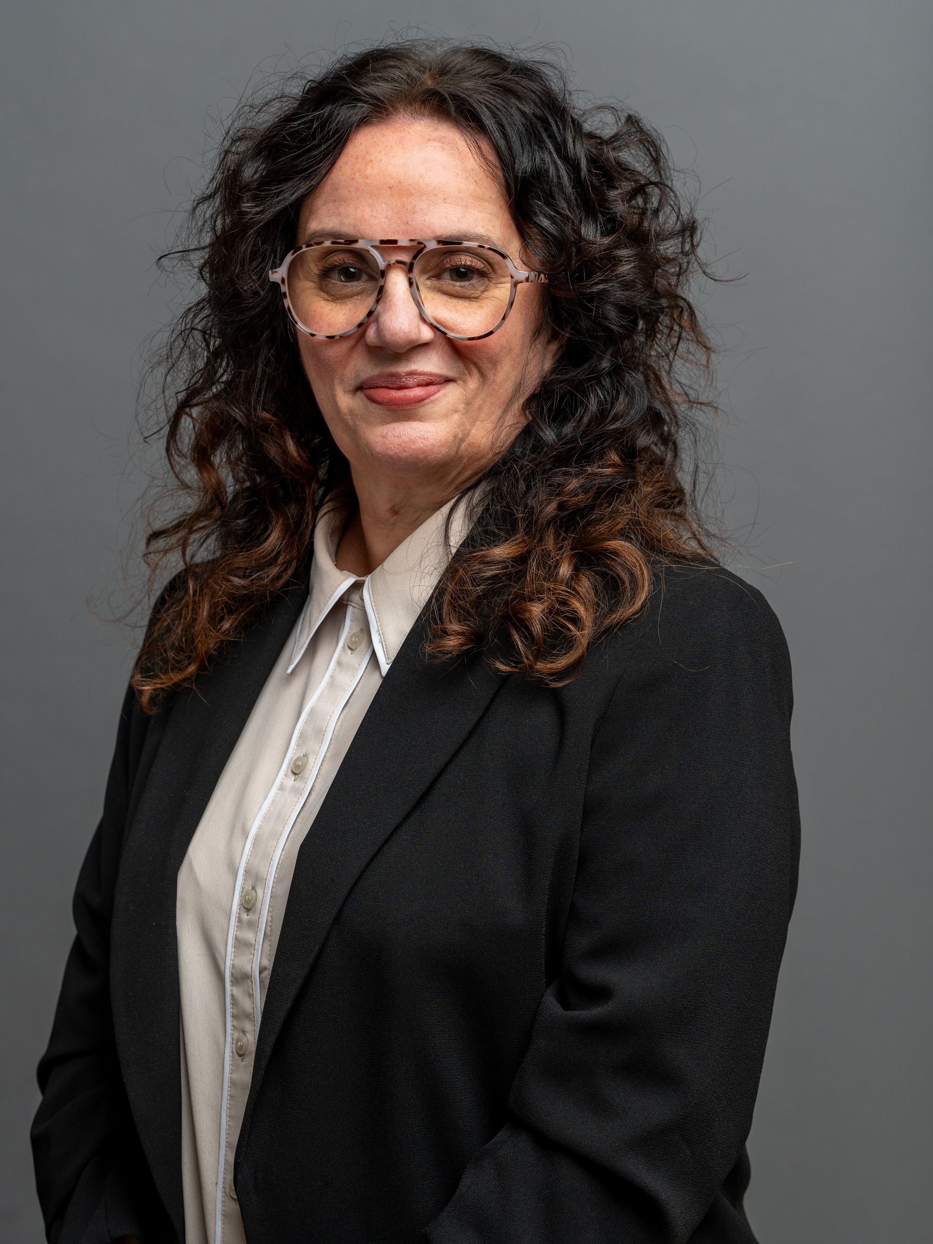 Woman with curly brown hair, glasses, wearing a blazer and button-up shirt, smiles at the camera.