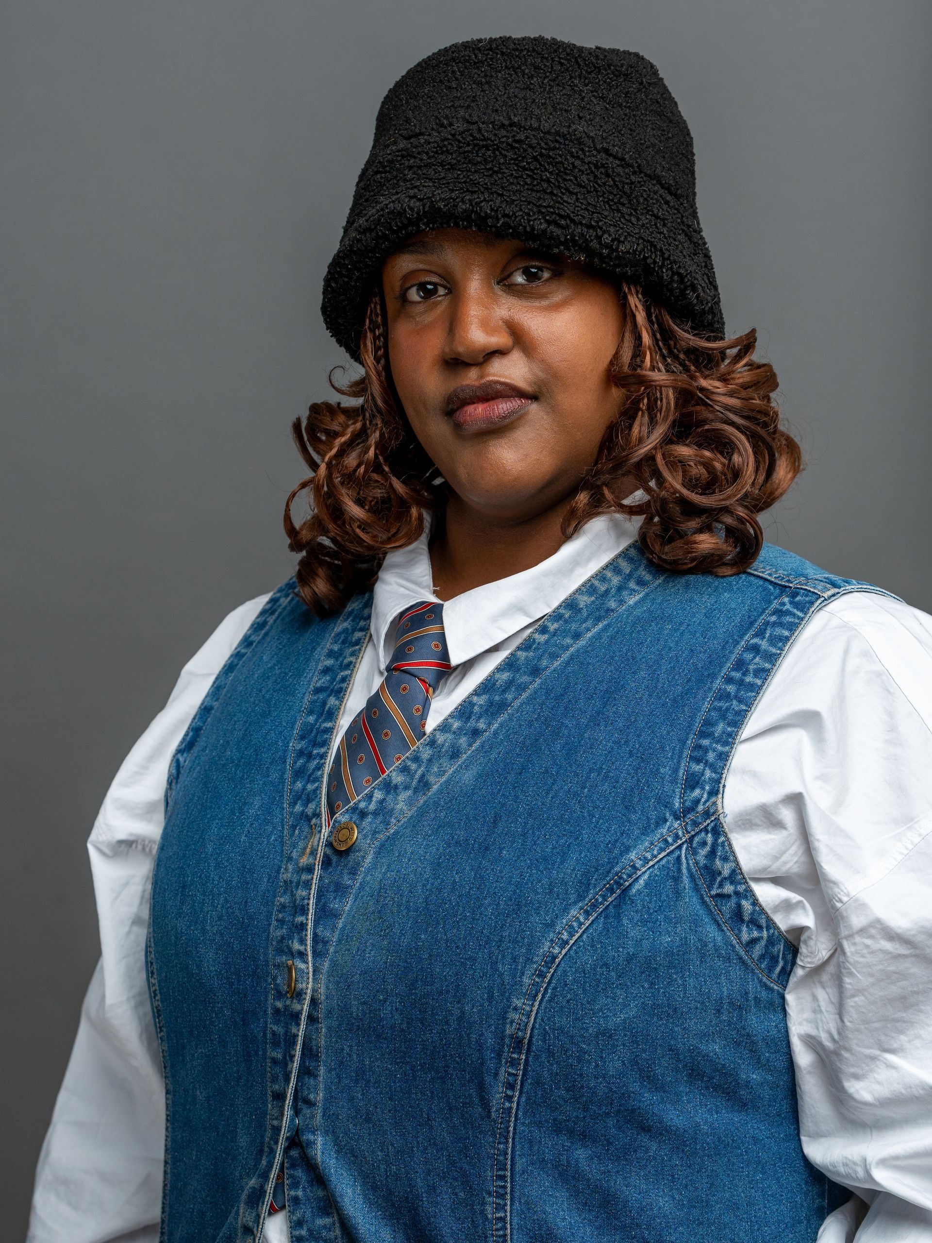 Person wearing a denim vest, patterned tie, and black fuzzy hat, with shoulder-length, curled brown hair.
