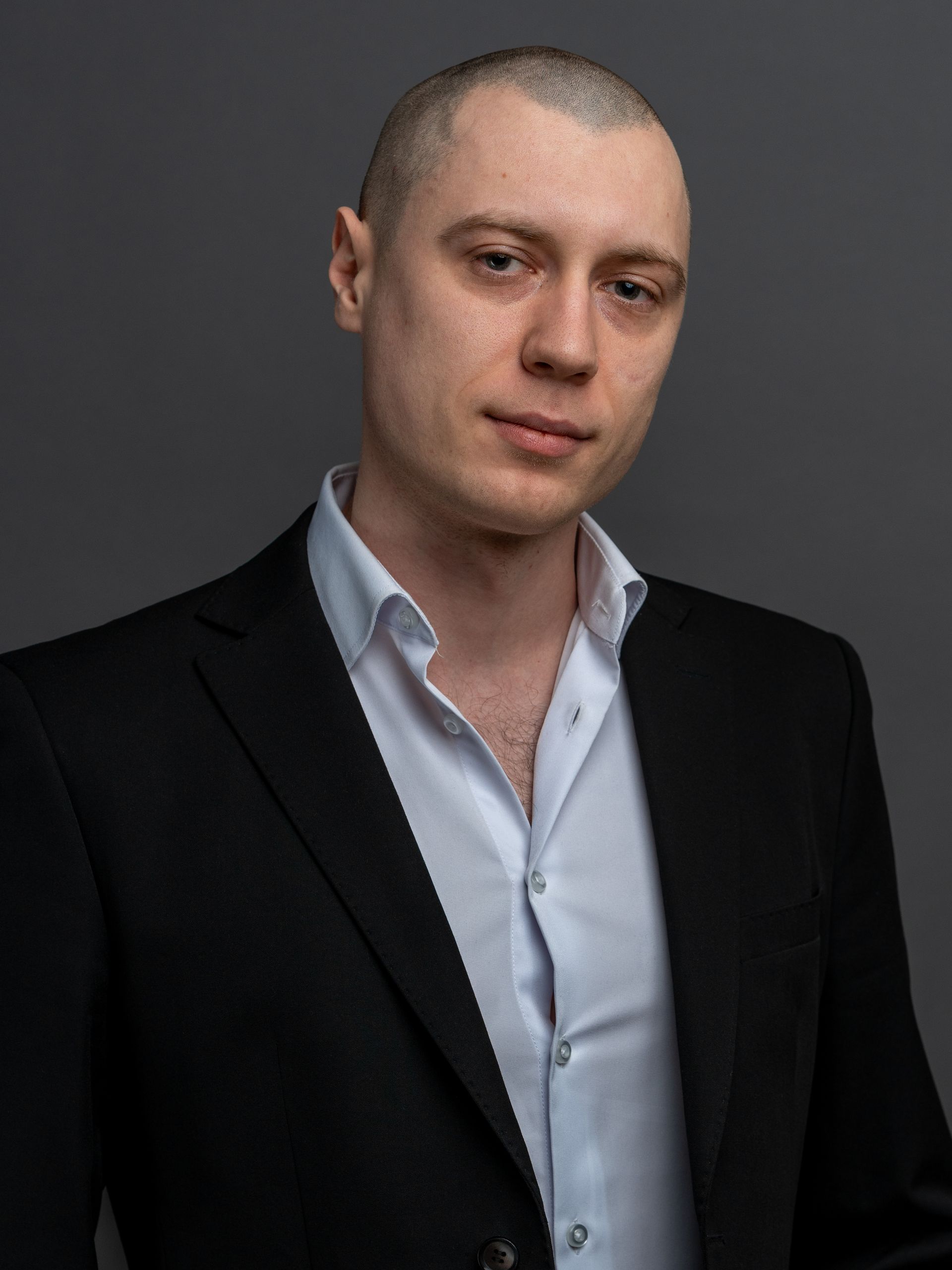 Man with shaved head wearing a black suit jacket and white shirt, looking at the camera.