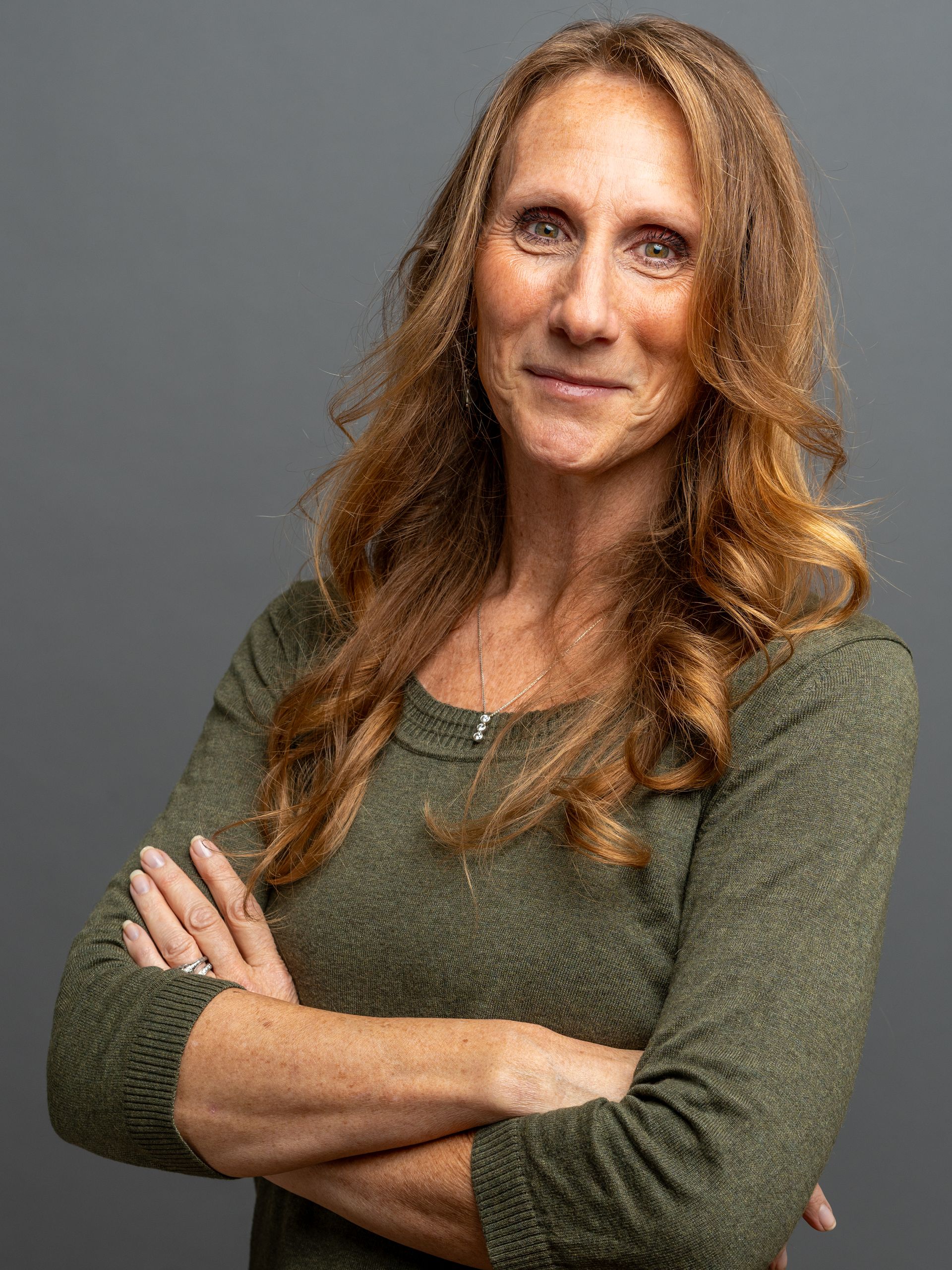 Woman with auburn hair and arms crossed, wearing a green long-sleeved shirt, on a grey background.
