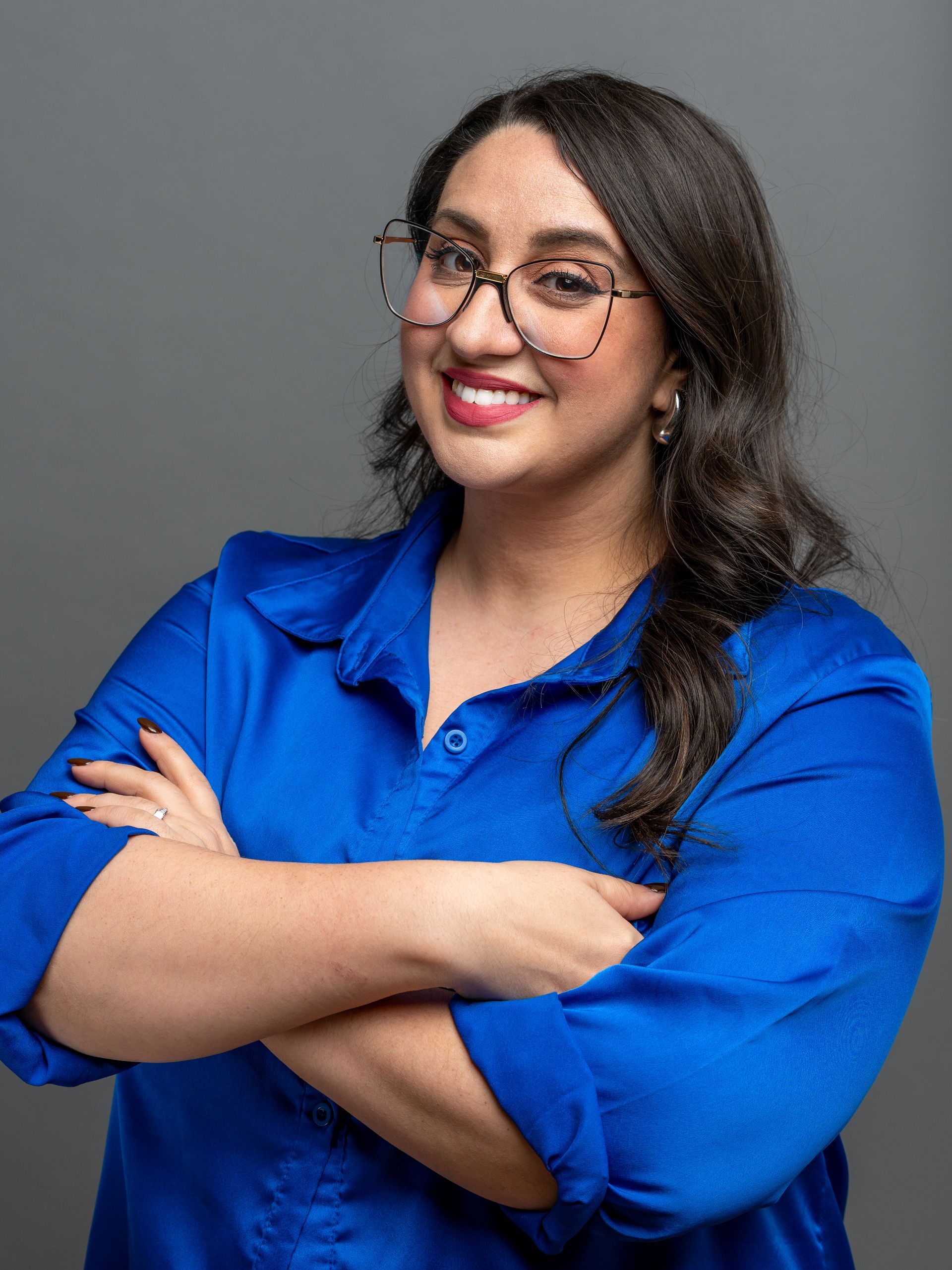 Woman with glasses and crossed arms, wearing a blue shirt, smiling against a gray background.