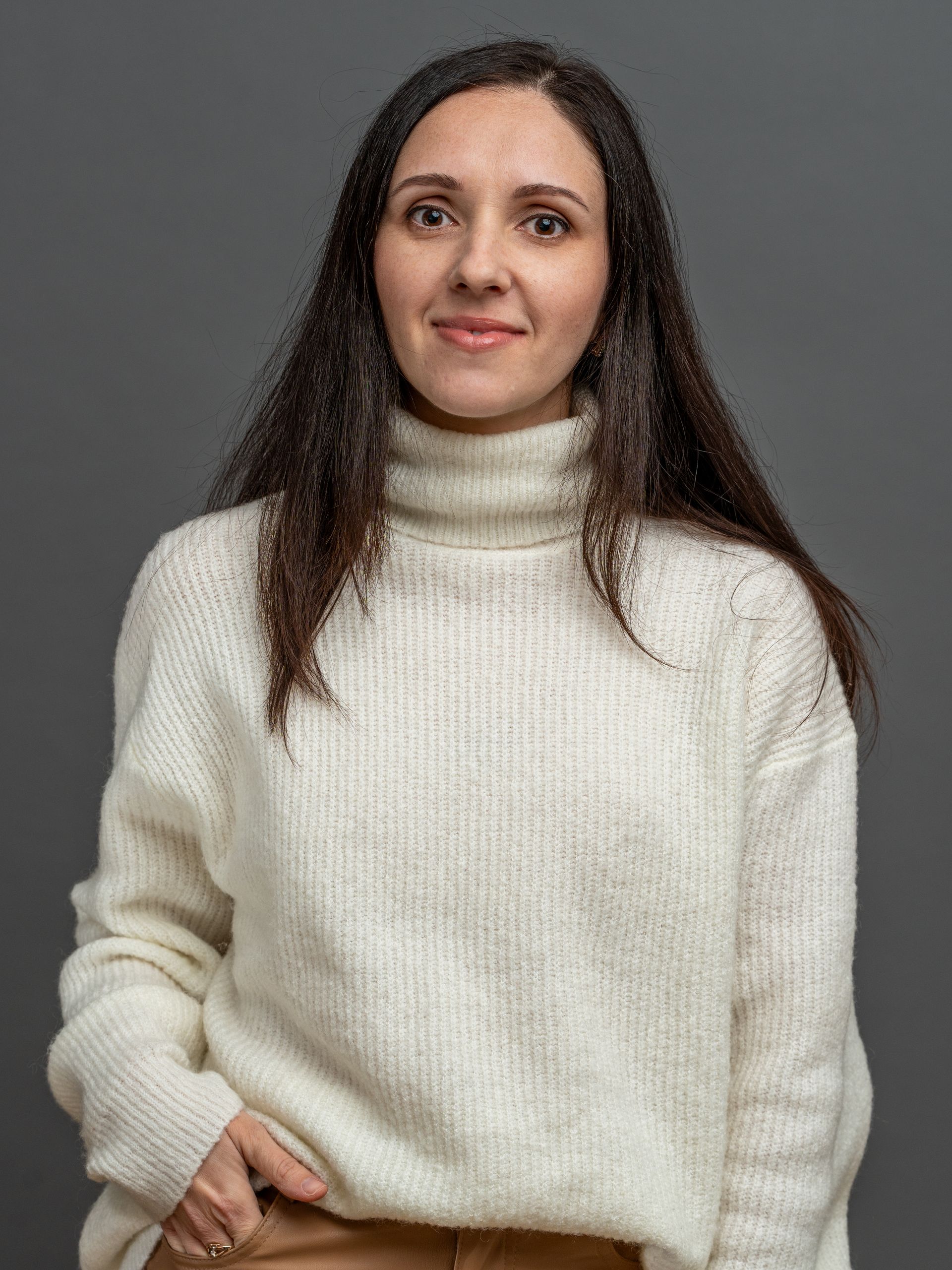 Woman in white turtleneck sweater, smiling, with long dark hair, against a gray background.