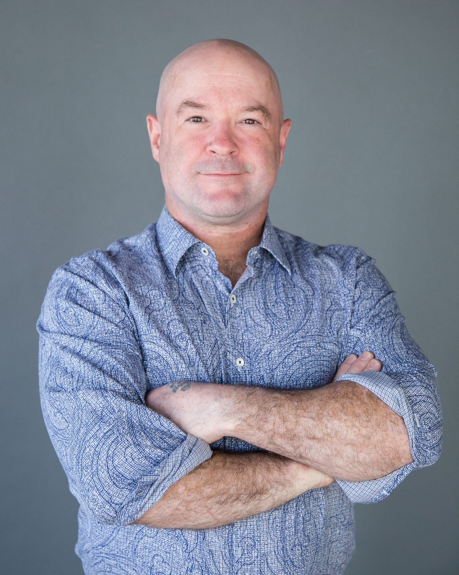 Bald man with arms crossed, wearing a blue paisley shirt, against a gray background.